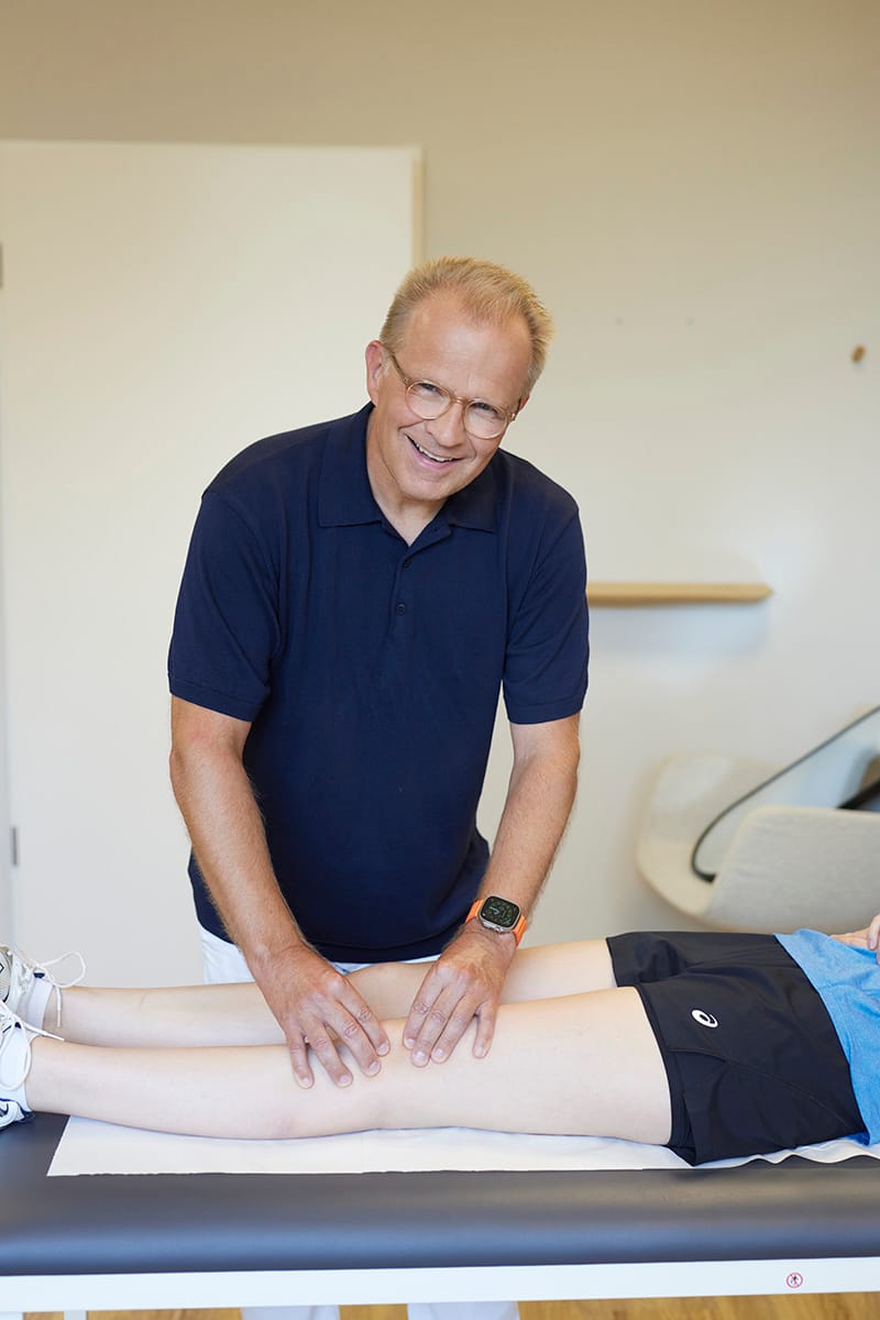 Prof. Dr. Oliver Tobolski tastet das Knie eines Patienten auf der Behandlungsliege mit beiden Händen ab