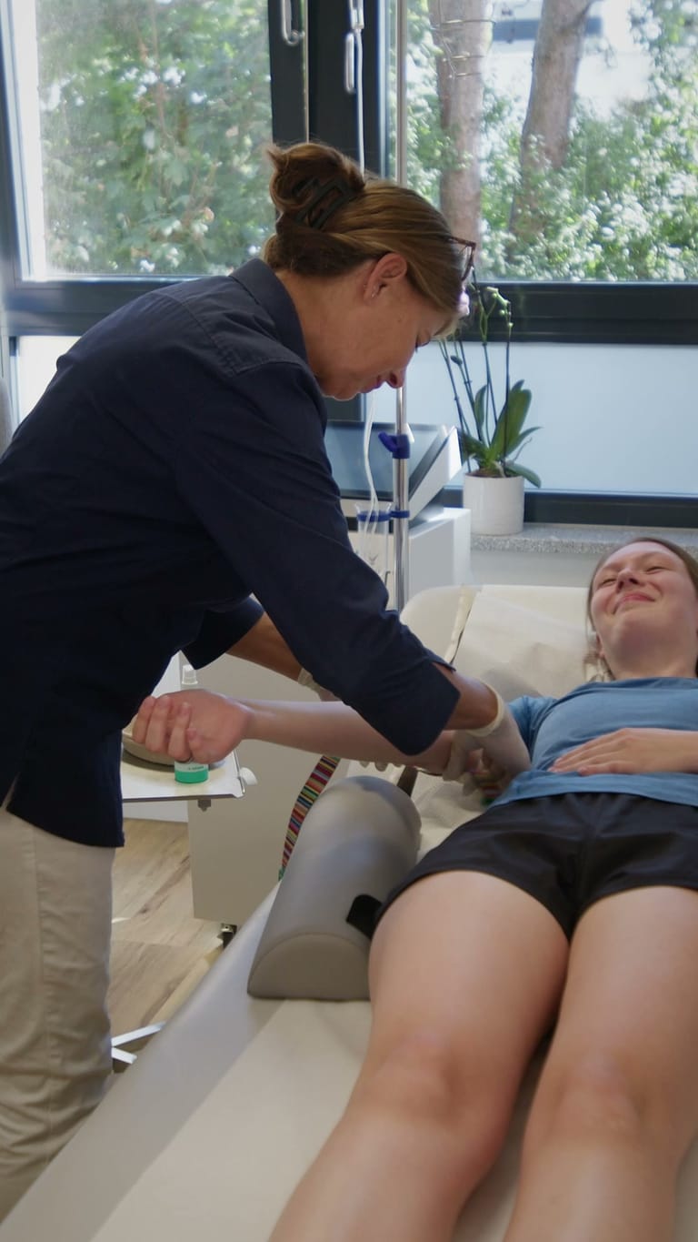 Mitarbeiterin legt einer Patientin auf der Liege den Zugang für die Infusion