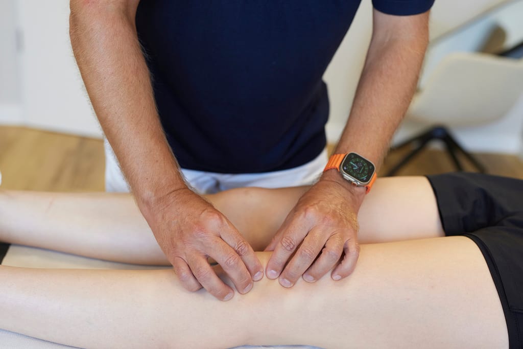 Nahaufnahme: Ein Arzt tastet mit beiden Händen die Gelenkspalte am Knie eines liegenden Patienten ab