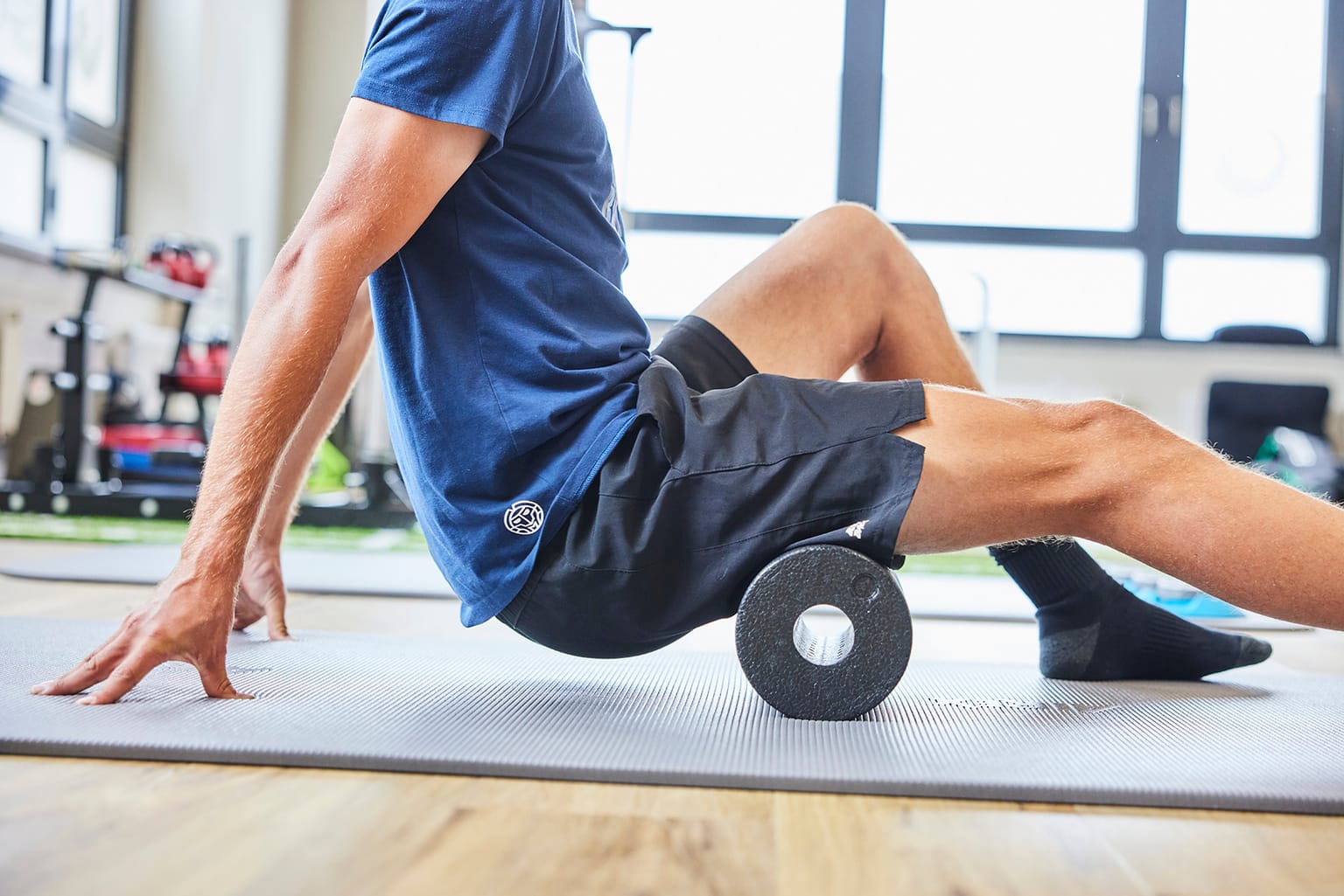 Mann in blauem T-Shirt rollt auf einer Gymnastikmatte die Rückseite des Oberschenkels mit einer schwarzen Faszienrolle aus