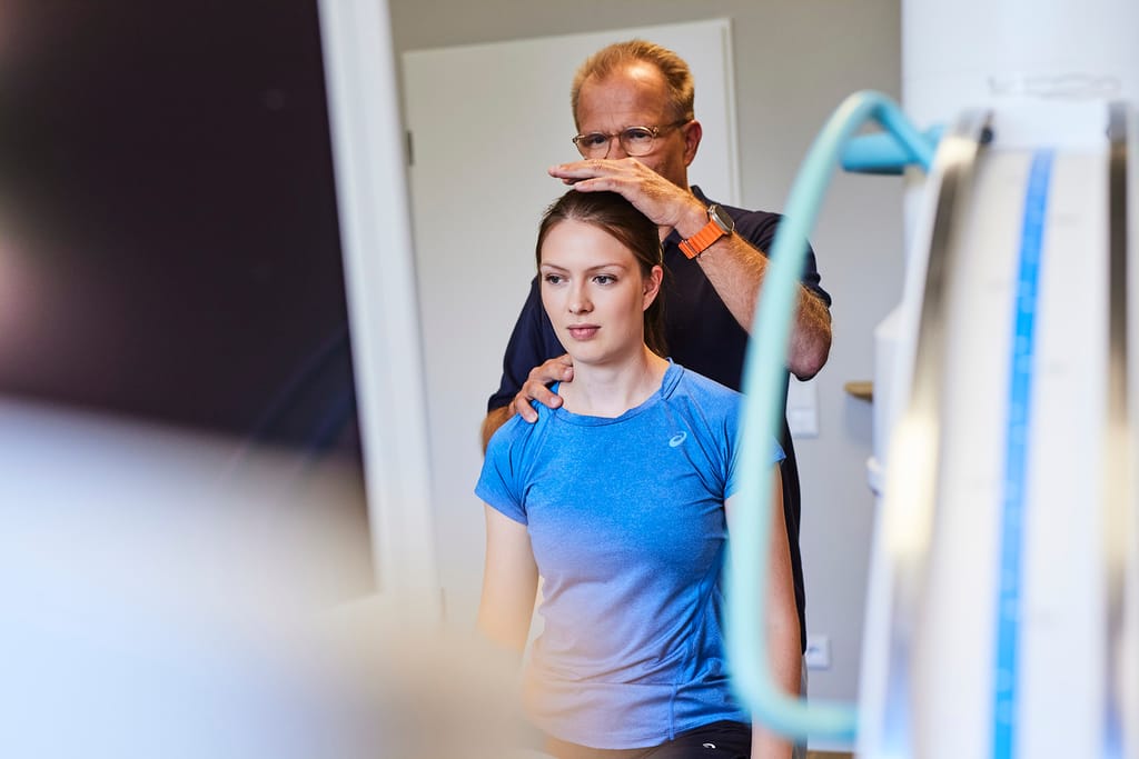 Ein Arzt prüft stehend die Beweglichkeit von Kopf und Wirbelsäule einer Patientin